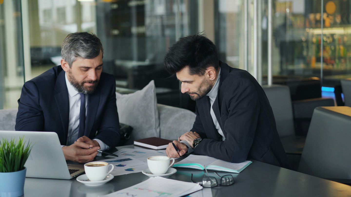 Two businessmen discuss financial charts at a table.
